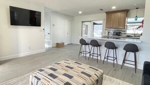 A modern kitchen with a bar area, a flat-screen TV mounted on the wall, and a patterned ottoman in the living area.