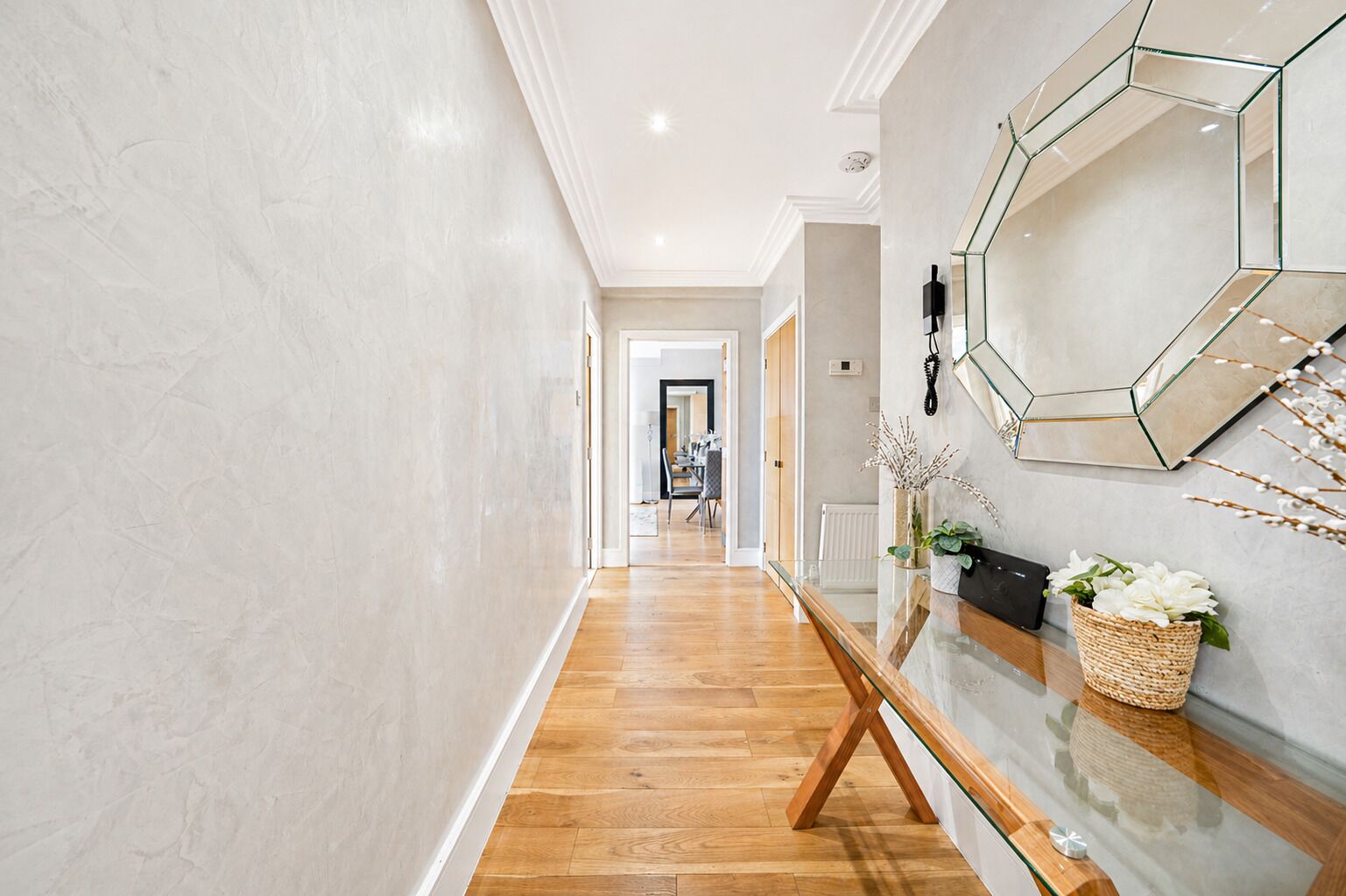 A hallway with a wooden floor, a glass table, and a wall-mounted mirror.