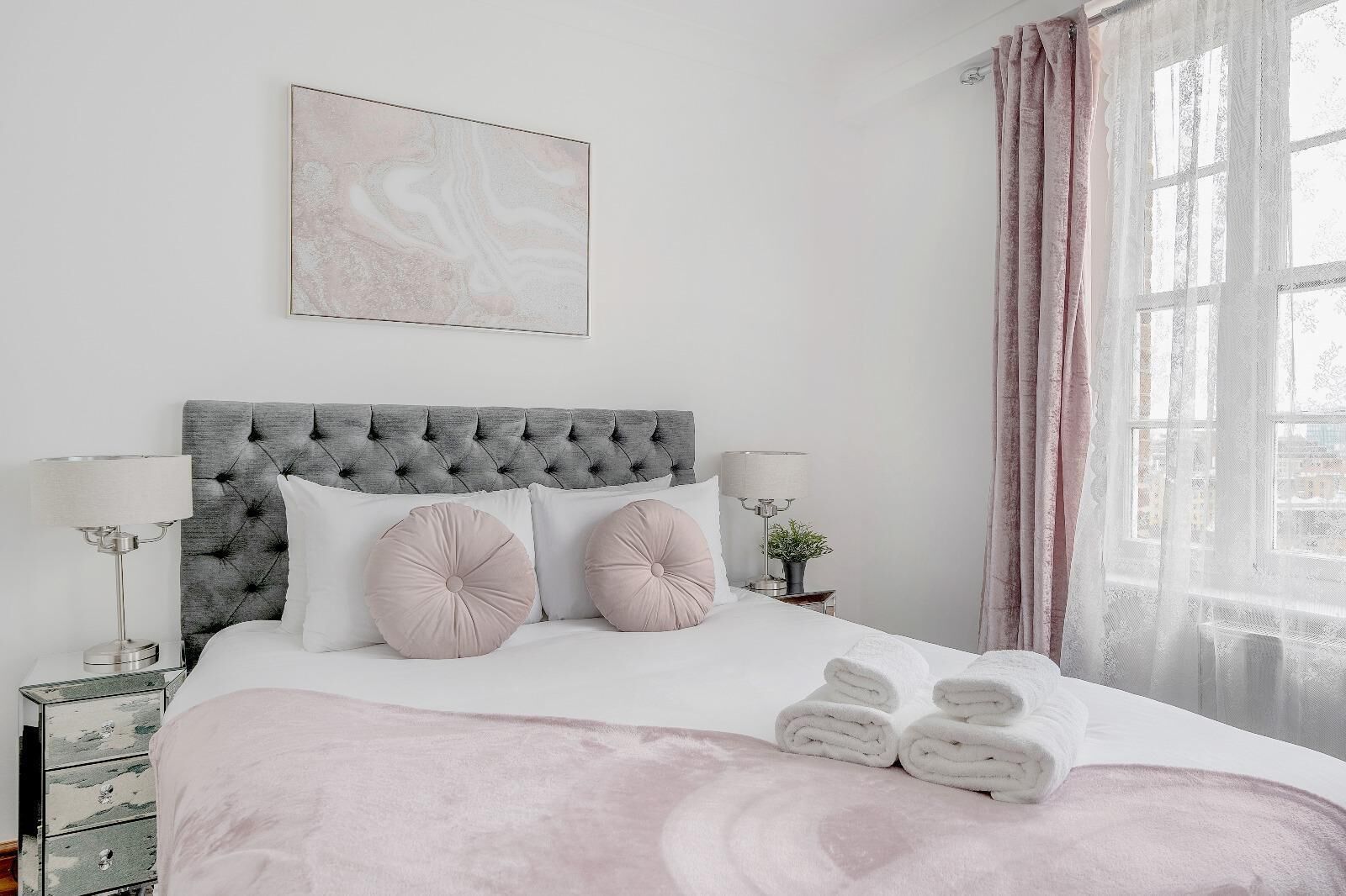 Un dormitorio con cabecera capitonada, dos lámparas de noche, cama con ropa blanca y almohadas rosas, ventana con cortinas traslúcidas y vista panorámica de la ciudad.