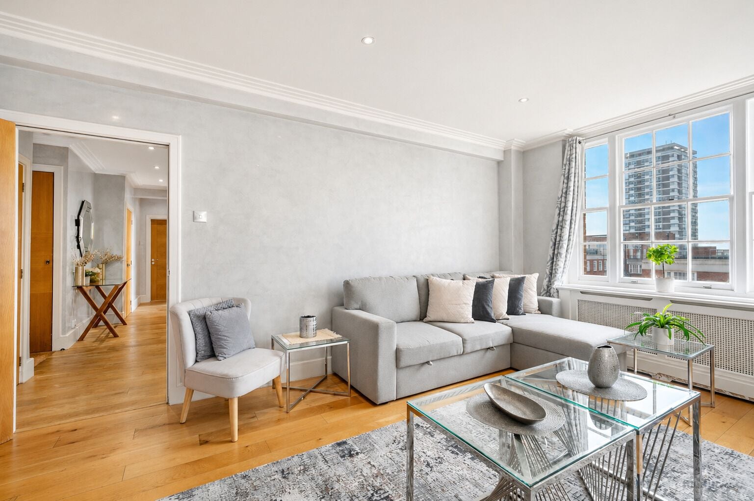 A modern living room with a grey sofa, a glass coffee table, and a view of buildings through large windows.