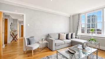A modern living room with a grey sofa, a glass coffee table, and a view of buildings through large windows.