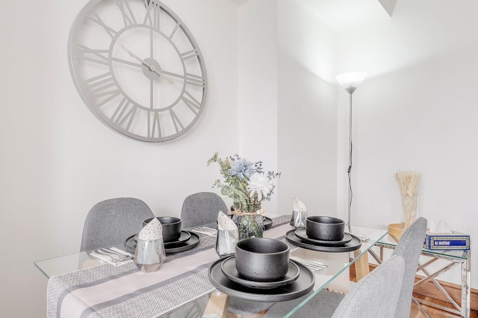 Et spiseområde med et glasbord, grå stole, et stort vægur og en vase med blomster.