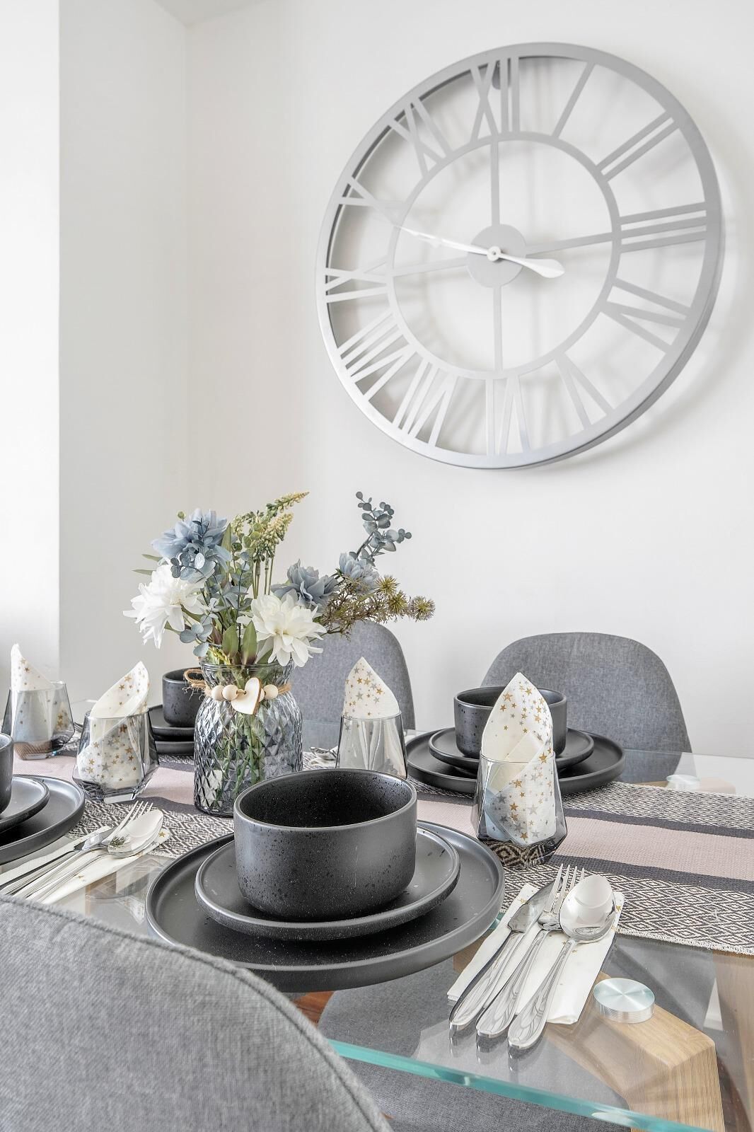 Une table à manger dressée avec des assiettes, des verres et des couverts, un vase rempli de fleurs et une grande horloge murale.