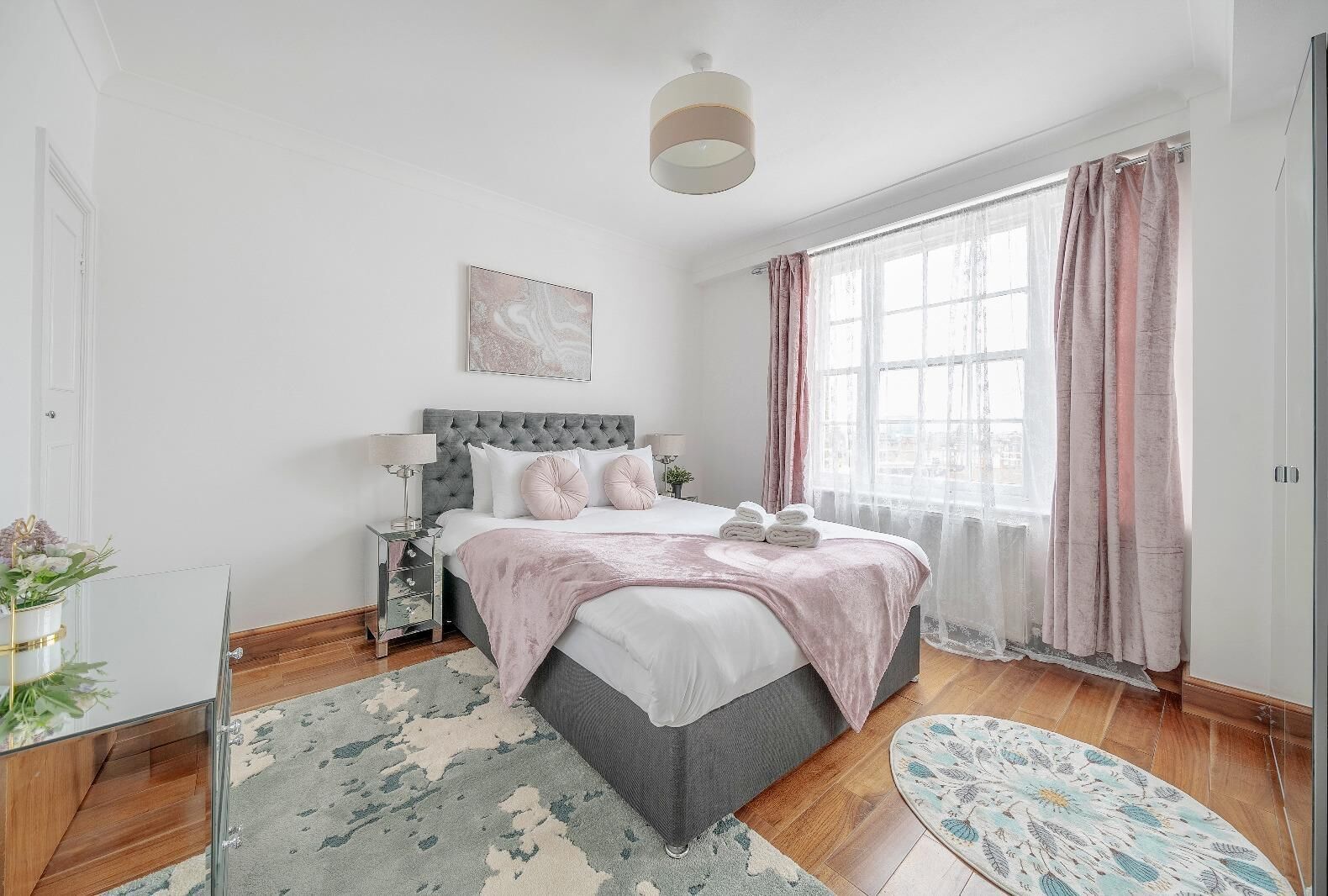 Un dormitorio con cama, mesitas de noche, una ventana con cortinas, un cuadro en la pared y vistas al exterior.