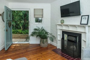 Une pièce dotée d’un plancher en bois, d’une cheminée blanche, d’un téléviseur fixé au-dessus et d’une porte donnant sur un jardin.