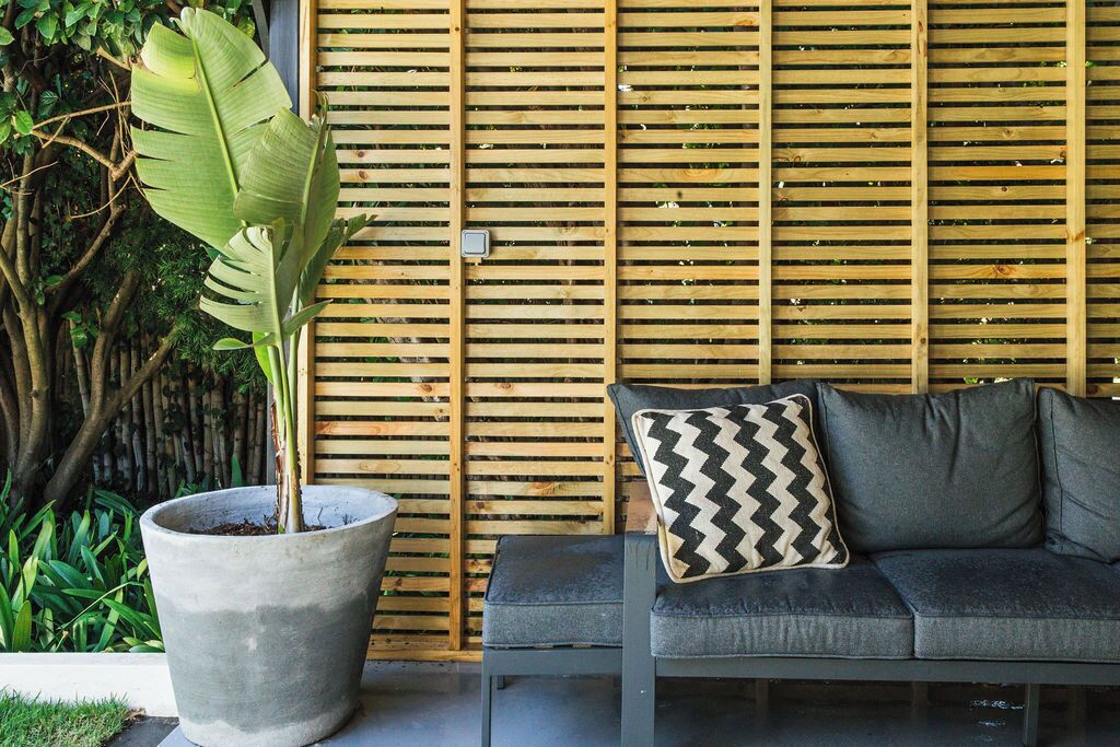 A modern outdoor seating area with a concrete planter, a grey sofa with a patterned pillow, and a wooden slatted wall.
