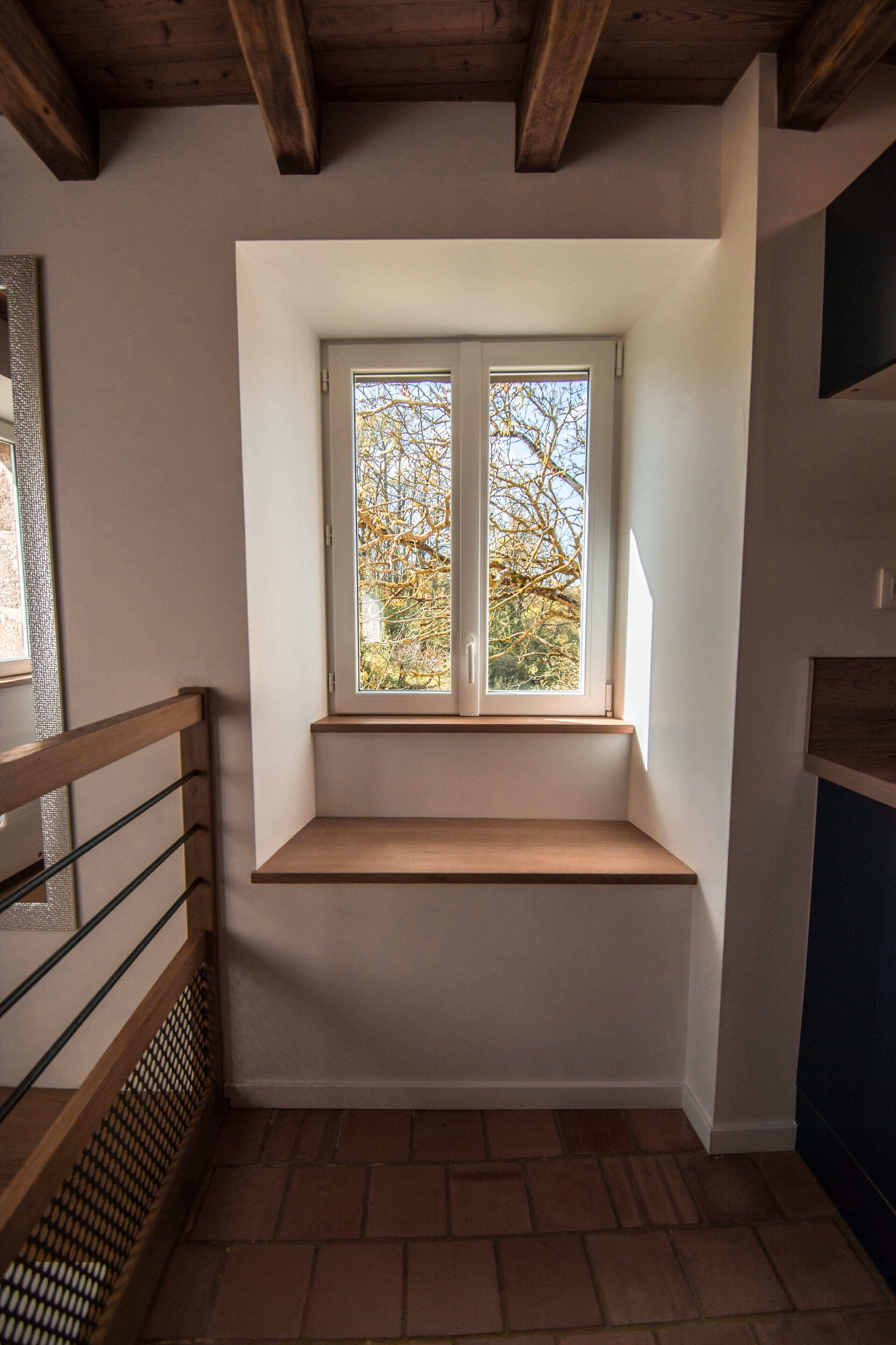 Una habitación de esquina con un banco empotrado bajo una ventana, una escalera de madera y piso de ladrillo.