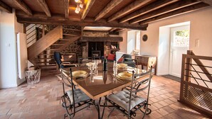 Ein Essbereich mit Holztisch, gedeckt für vier Personen, umgeben von gepolsterten Stühlen, einem Steinkamin und einer Treppe, die zu einer weiteren Ebene führt.