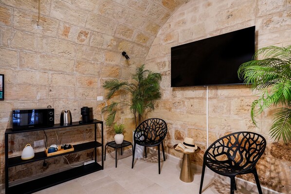 A stone-walled room with a flat-screen TV, a coffee maker, a microwave, a small table with a plant, and two chairs.