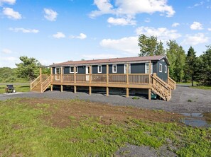 A two-story cabin with a wooden deck and a small porch, surrounded by a gravel driveway and grassy area.