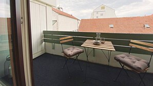 Ein Balkon mit Tisch und zwei gepolsterten Stühlen, einem Fenster und Blick auf Gebäude mit roten Dächern.