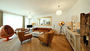 A modern living room with a brown sofa, two armchairs, a coffee table, and a unique round chair. There is a large mirror on the wall, a floor lamp, and a sideboard with decorative items. - Appartement fur 5 Personen in Strausberg (Strausberg)