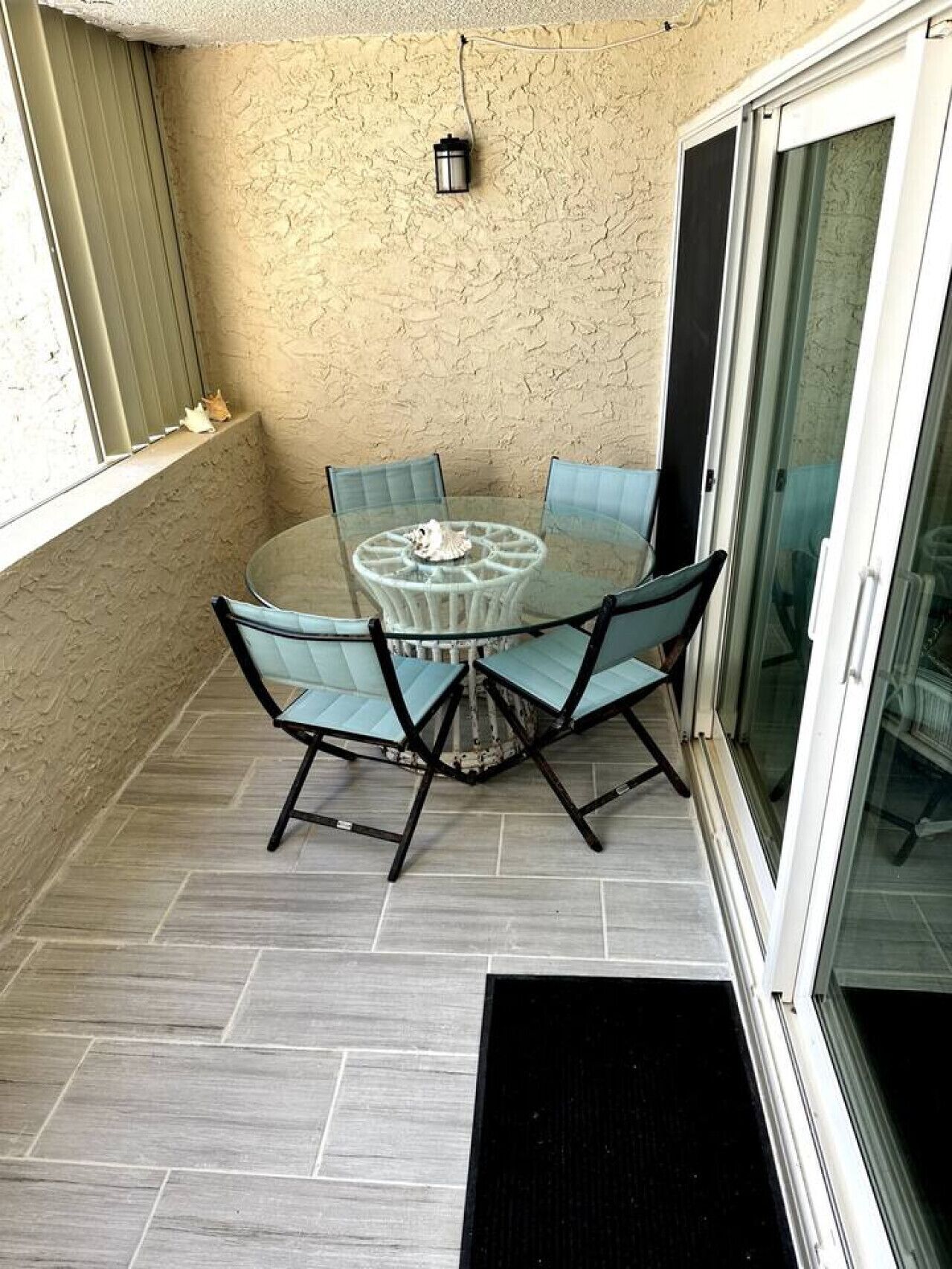 Une petite terrasse extérieure avec une table en verre et quatre chaises, un petit luminaire fixé au mur et une porte-fenêtre coulissante.