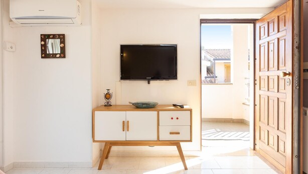 A room with a flat-screen TV mounted on the wall, a wooden cabinet with drawers, a small decorative item on the cabinet, and a door leading outside.