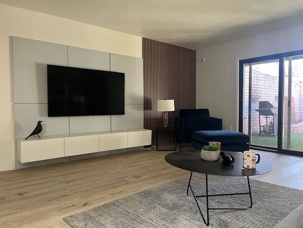 A modern living room with a flat-screen TV mounted on a white wall, a dark blue sofa, a small round coffee table with a plant and mug, and a sliding glass door leading to a backyard. - Walk to Spring Training - 3BR home in Scottsdale (Scottsdale)