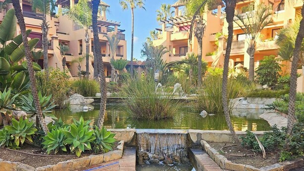A courtyard with a small water feature, surrounded by palm trees and a brick pathway. - Beachfront Apartment in San Pedro - Pass the Keys (Marbella)