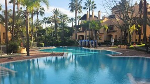 A large, clear swimming pool surrounded by palm trees and a building with a tiled roof. - Beachfront Apartment in San Pedro - Pass the Keys (Marbella)