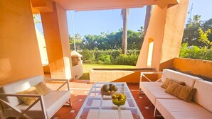 A sunlit patio with a glass-top table, white sofas, and a view of a lush garden. - Beachfront Apartment in San Pedro - Pass the Keys (Marbella)