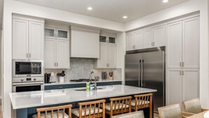 A modern kitchen with white cabinets, a stainless steel refrigerator, a microwave, and a large island with bar stools.