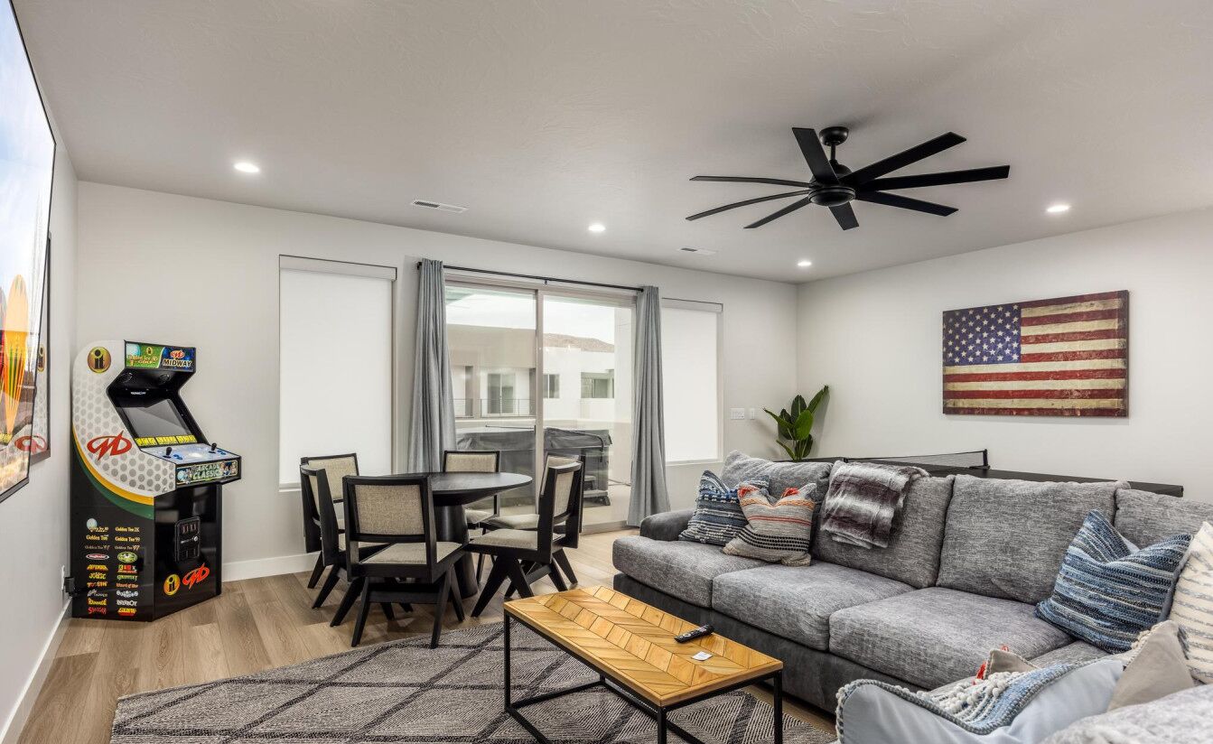 A modern living room with a grey sofa, a coffee table, and a ceiling fan. There is a dining area with a table and chairs, and a jukebox on the left. An American flag is on the wall.