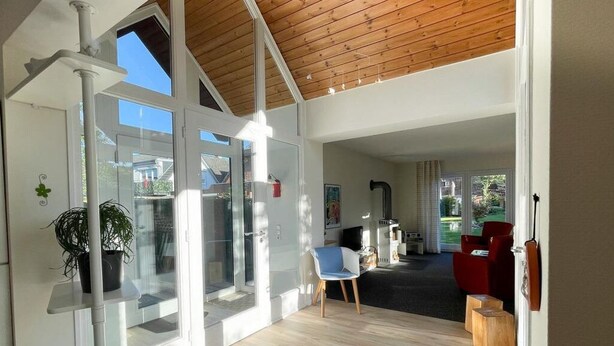 Un interior moderno con entrada de puerta de vidrio, techo de madera, zona de estar con una silla roja y una planta en maceta.
