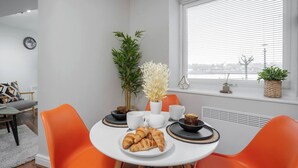 A dining area with a round table, two orange chairs, a white plate with croissants, and a window with blinds.
