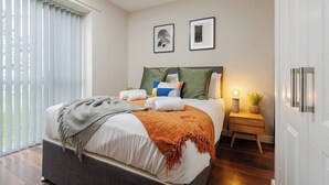A modern bedroom with a large bed, a bedside table with a lamp, and a view through a window with blinds.