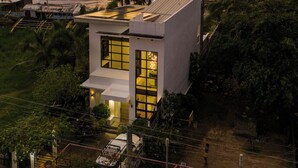 A modern house with a large glass window, surrounded by greenery and a view of the sea at sunset. - Ocean View (Naval)