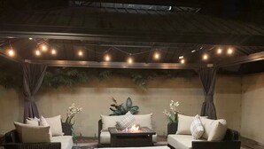 An outdoor patio with wicker furniture, a central fire pit, and string lights.