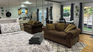 A modern bedroom with a large bed, two sofas, a desk, and a kitchen area visible through a glass partition.