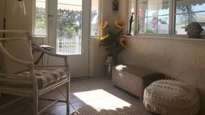 Una habitación acogedora con una silla de mimbre, un pequeño banco y una ventana que da a una cerca blanca y vegetación.