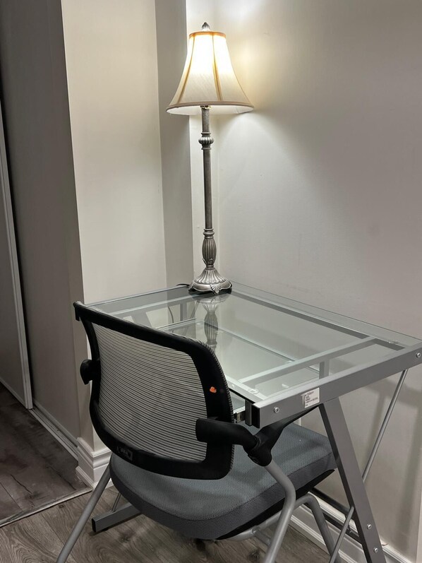 A glass-top table with a lamp, a black mesh office chair, and a white wall. - Comfy Basement Apartment with Private entrance and free car (Mississauga)