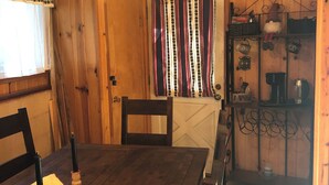 A wooden interior with a dining table, chairs, a window with curtains, and a shelf with kitchen items. - Charming Ski cabin with WiFi near Dodge Ridge, great for family or small group. (Mi-Wuk Village)