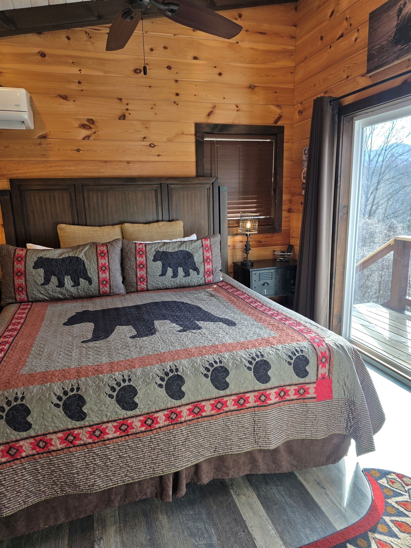 Ein Schlafzimmer mit Holzbalkendecke, einem Bett mit einer Bären-Quiltdecke, einem Nachttisch und einem Fenster mit Blick auf Bäume.