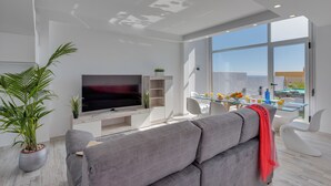 Een moderne woonkamer met een flatscreen televisie, een grijze bank en een eethoek met een glazen tafel en stoelen.