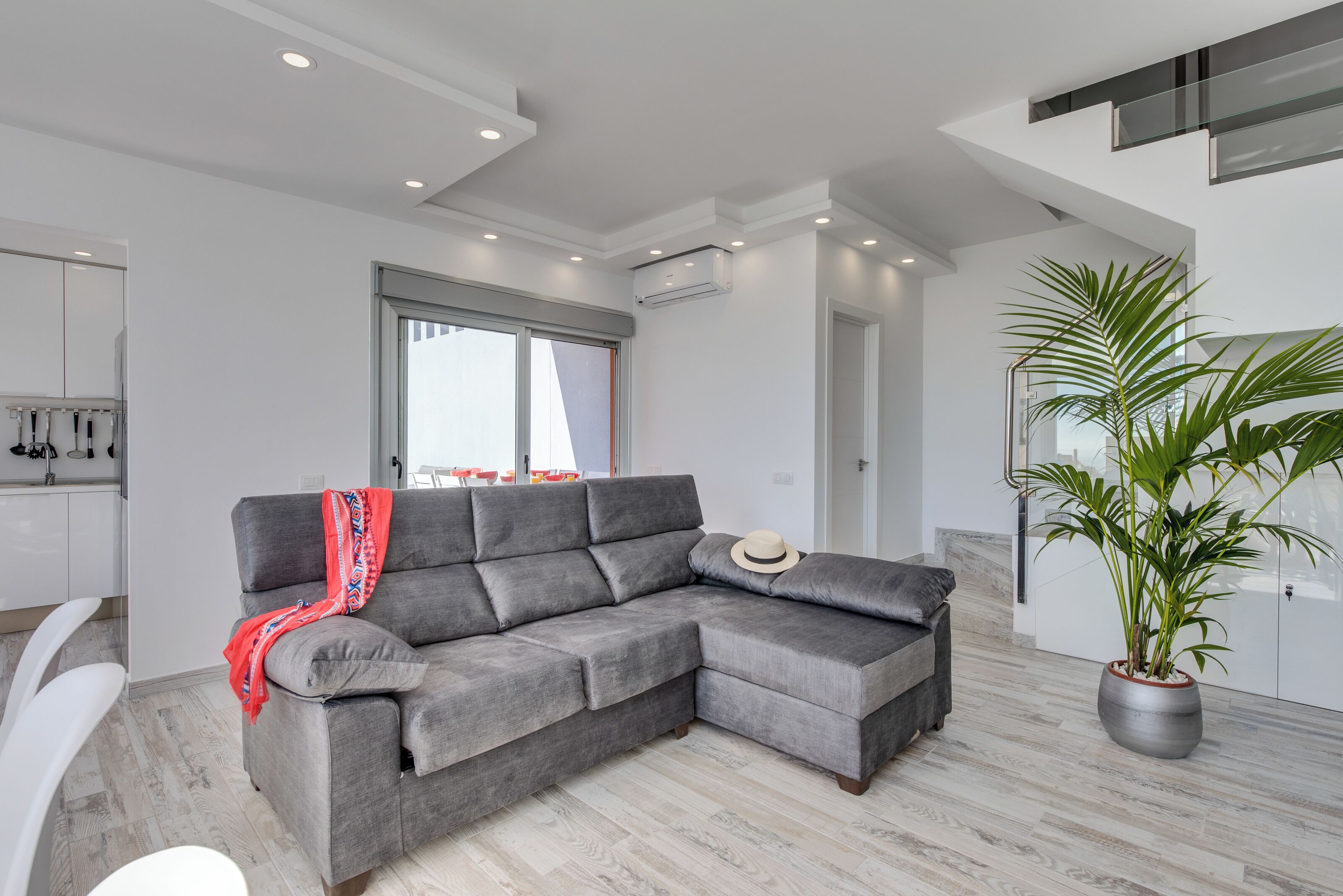 A modern living room with a grey sectional sofa, a large potted plant, and a view of the city through a window.