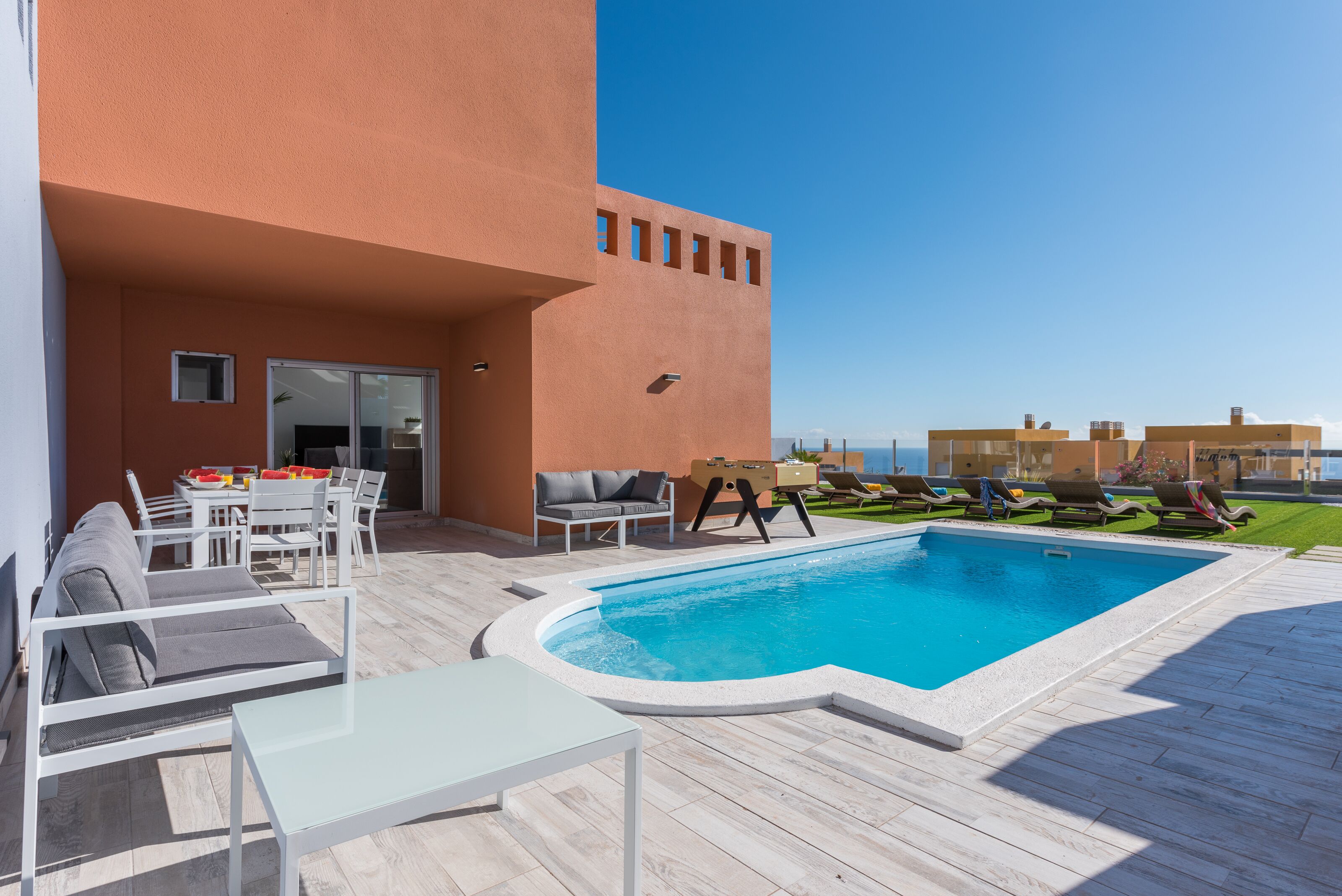 An outdoor swimming pool area with lounge chairs and a table, surrounded by a building with a flat roof and a clear sky.