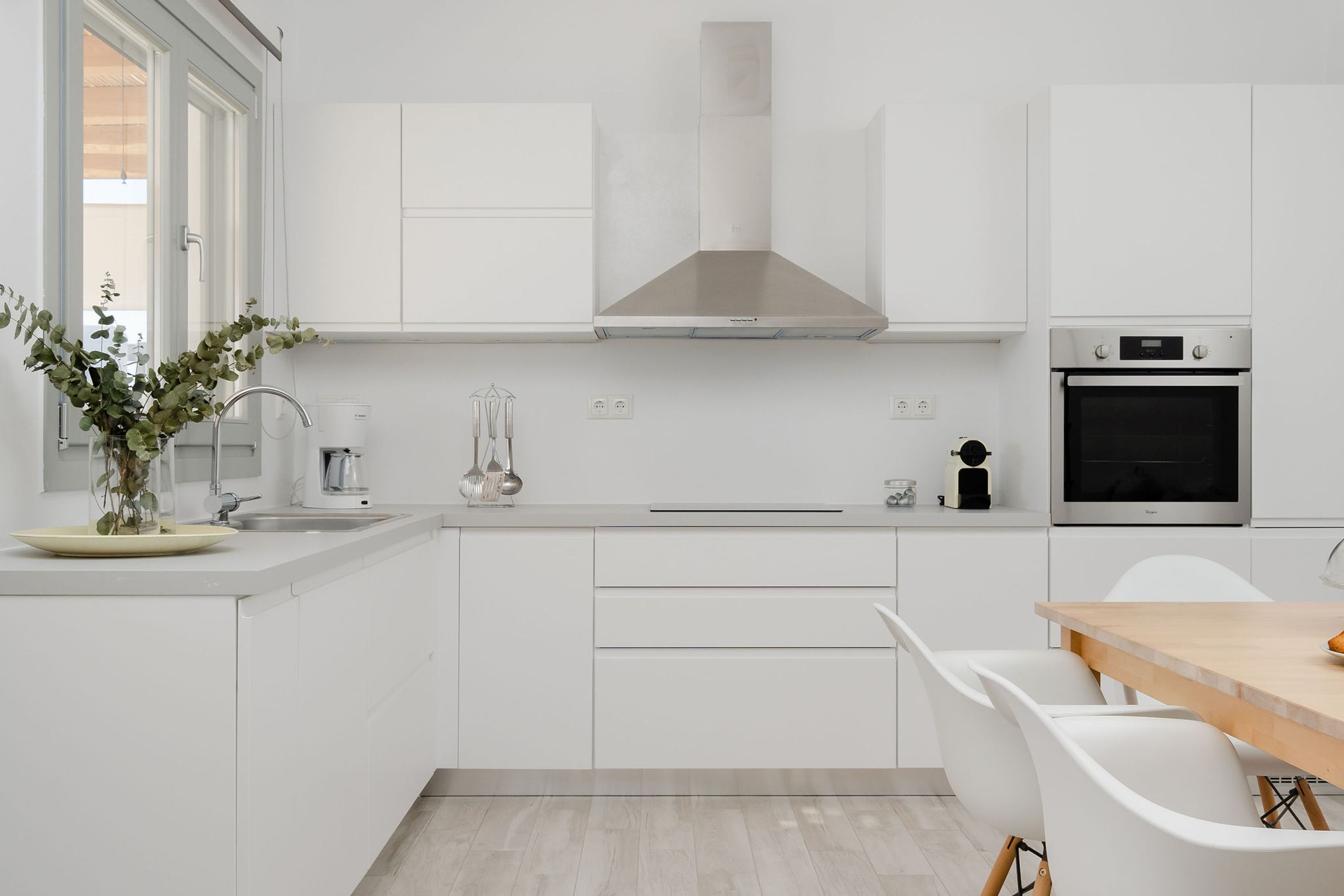 A modern kitchen with white cabinetry, a stainless steel range hood, a built-in oven, and a dining area with white chairs.