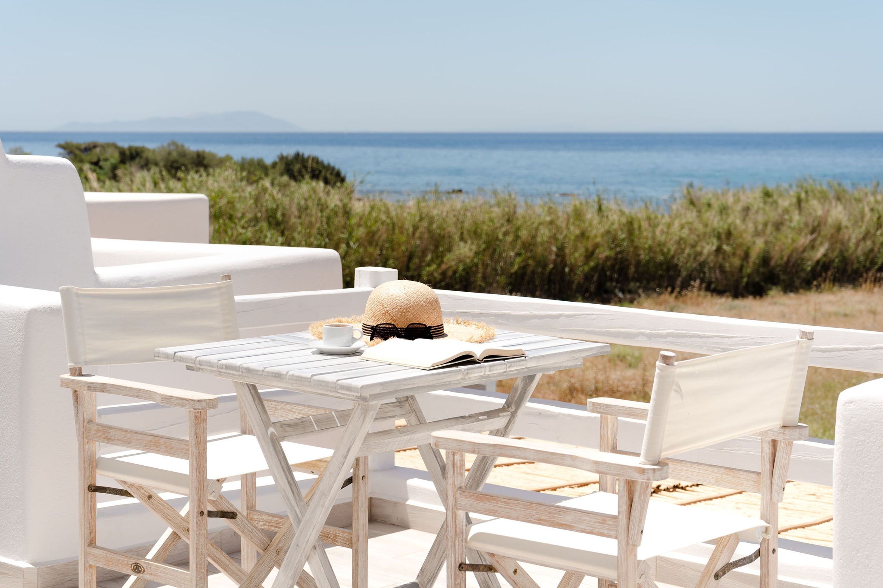 A white outdoor seating area with a table and chairs, overlooking a coastal landscape with vegetation and the sea.