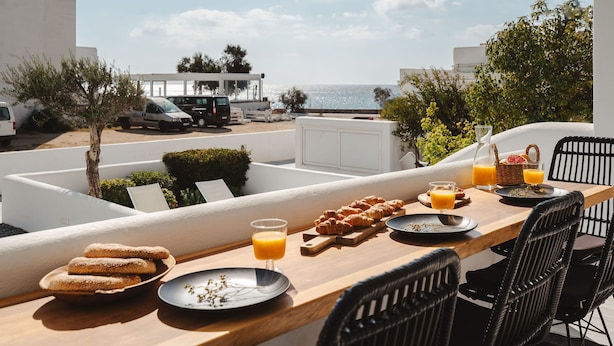Et morgenbord med croissanter, appelsinjuice og en kurv med jordbær, med udsigt over en strand og parkerede biler.