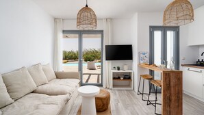A modern living room with a sofa, a coffee table, a TV, and a kitchen area with stools. - Villa Isola Plaka Naxos (Naxos)