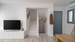 A modern living room with a flat-screen TV on a white console, a staircase leading to another room, and a wooden dining table. - Villa Kaya Plaka With 3 Bedrooms and 3 Bathrooms (Naxos)