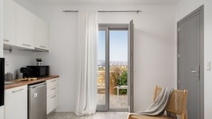 A compact kitchen with white cabinets, a dishwasher, and a microwave. A sliding glass door leads to a balcony with a wicker chair and a table. - Julie s Luxury Apartment (Naxos)