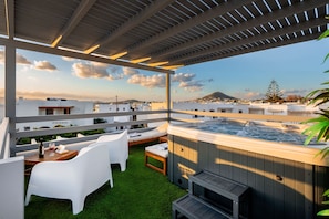 A rooftop terrace with a hot tub, white lounge chairs, and a wooden table with a bottle and glass.