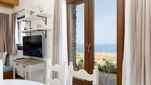 A room with a TV, white furniture, a view of the sea, and a stone wall.