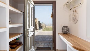 A narrow hallway with a door leading to an outdoor area with a view of stone ruins and a clear sky. Shelves line the left wall, and a wooden counter is on the right.