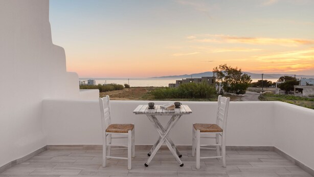 A balcony with two white chairs and a small table, overlooking a coastal landscape during sunset.