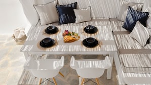 A white dining table with black bowls and a fruit basket, surrounded by white chairs and a sofa with patterned cushions.