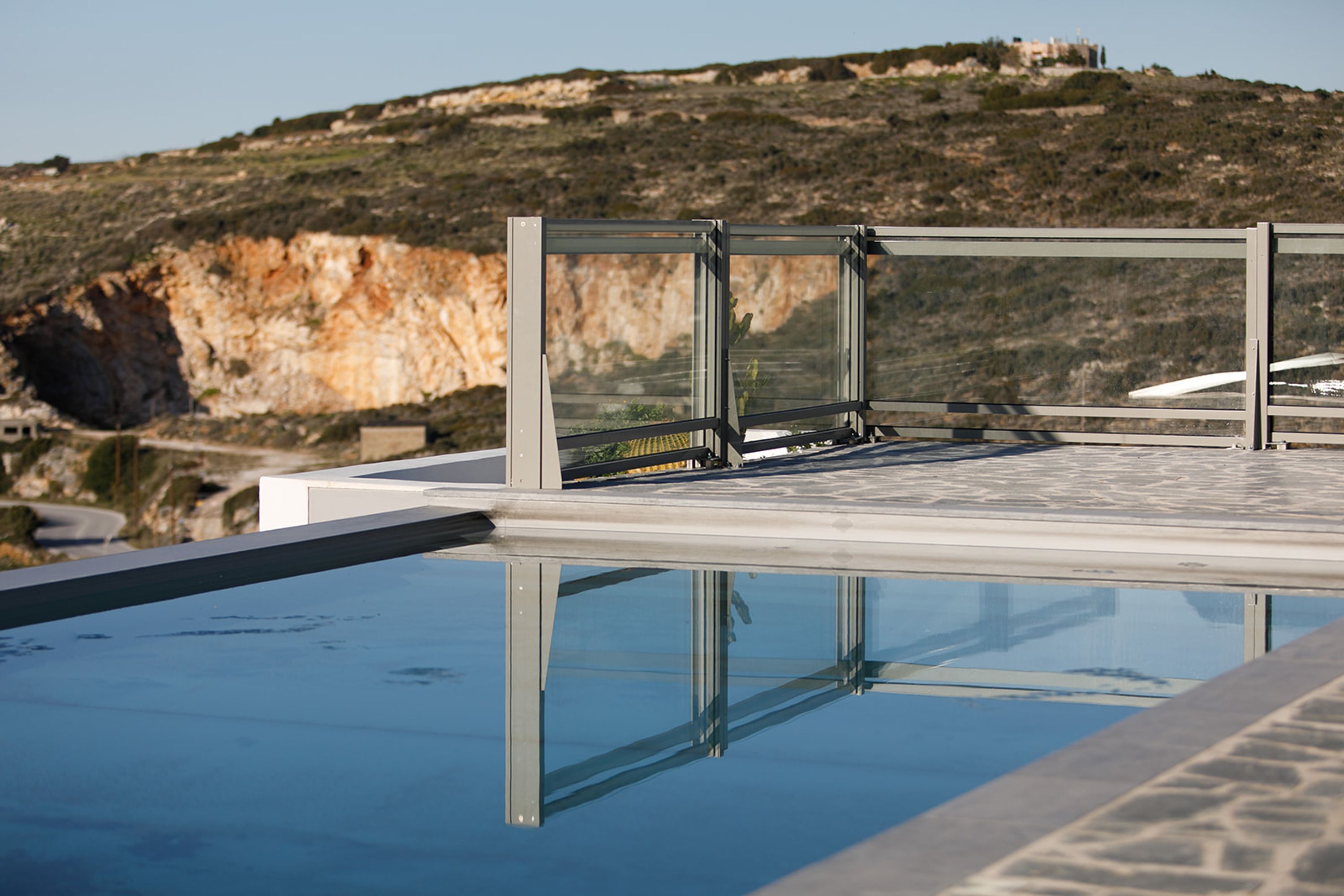 A modern infinity pool with clear water reflecting the surrounding landscape, including a rocky hillside and a distant building.