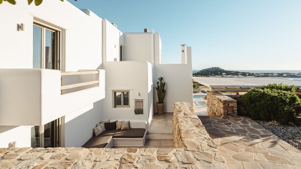 Un bâtiment moderne aux murs blancs, avec un chemin de pierre menant à un espace aménagé pour s’asseoir, d’où l’on a une vue sur une plage et l’océan.
